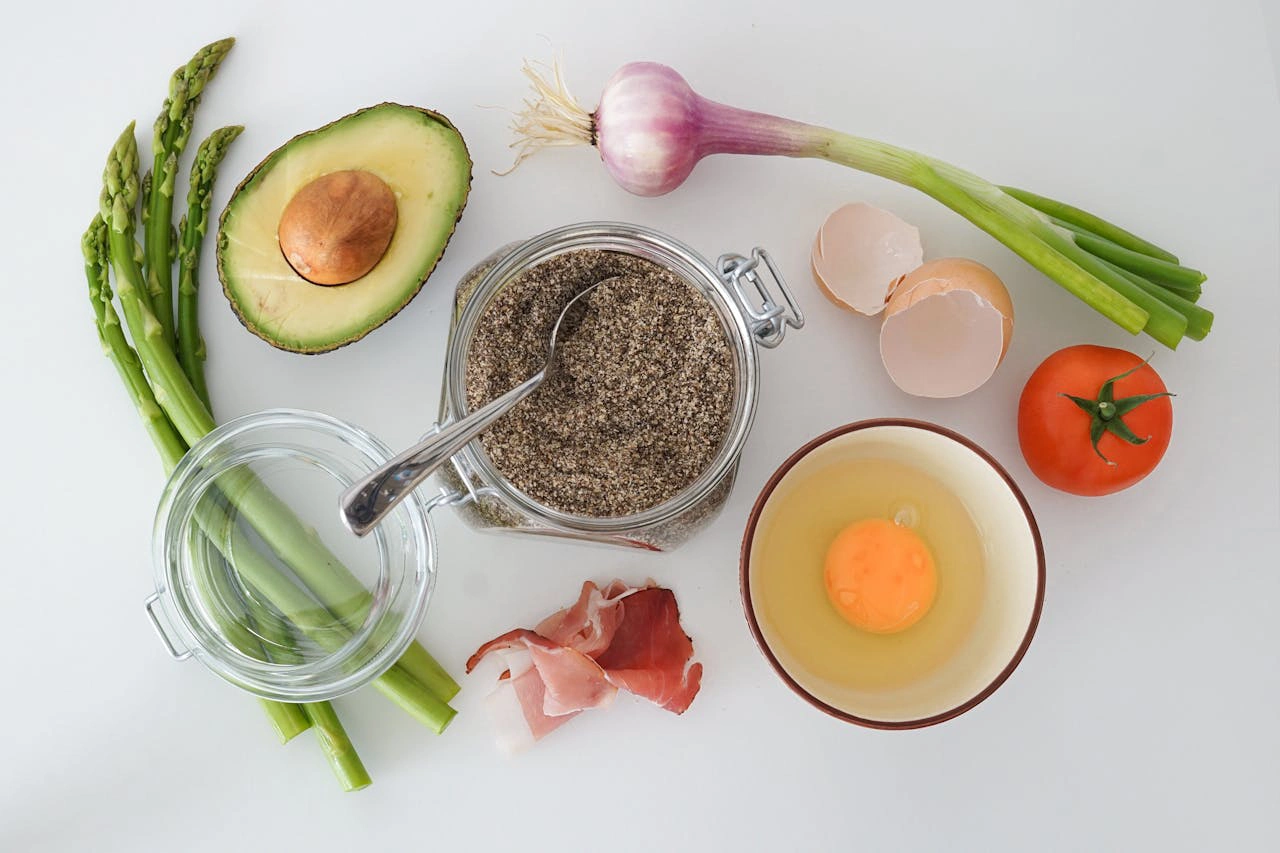 Fresh ingredients including avocado, asparagus, egg, tomato, onion, chia seeds, and bacon on a white table.